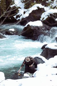 A man in winter clothing gathers water from a flowing river in a snowy landscape.
